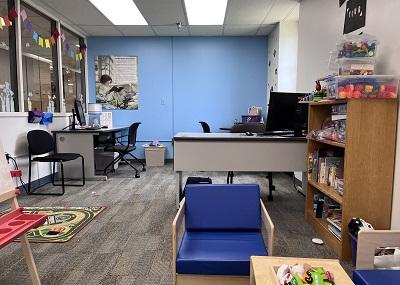 A carpeted room with two desks with monitors, blue chairs around a low table, and shelves of tabletop games.