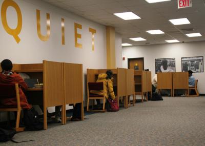 Students sitting at a row of individual partitioned study desks along a wall that has Quiet painted on it. The area is carpeted and has overhead lighting.
