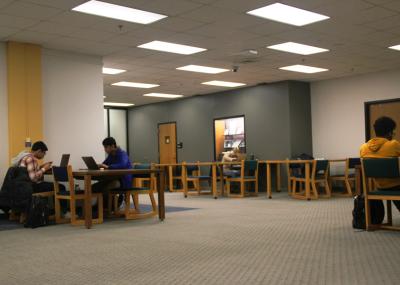 Carpeted room with overhead lighting featuring tables arranged with cushioned desk chairs around them. Students are working alone or in pairs at the tables.