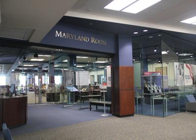 Floor to ceiling windows with a view of an exhibition space and reception desk in the foreground. A person is leaned over a table in an open space area in the background. A sign that reads Maryland Room runs across the top of the entrance.
