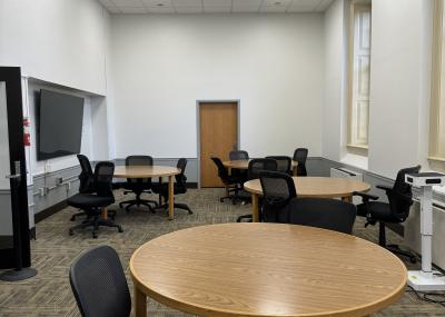 Three circular tables surrounded by wheeled armless chairs with mesh backs in a spacious room with tall window and a large monitor. Not pictured: a second large monitor and two wheeled whiteboards.